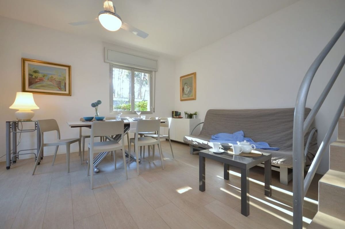 A modern living room is shown with a double sofa-bed and a wooden coffee table. A dining table with four chairs is set near a window, which allows natural light to brighten the space. Wall art decorates the walls, and a ceiling fan is visible.