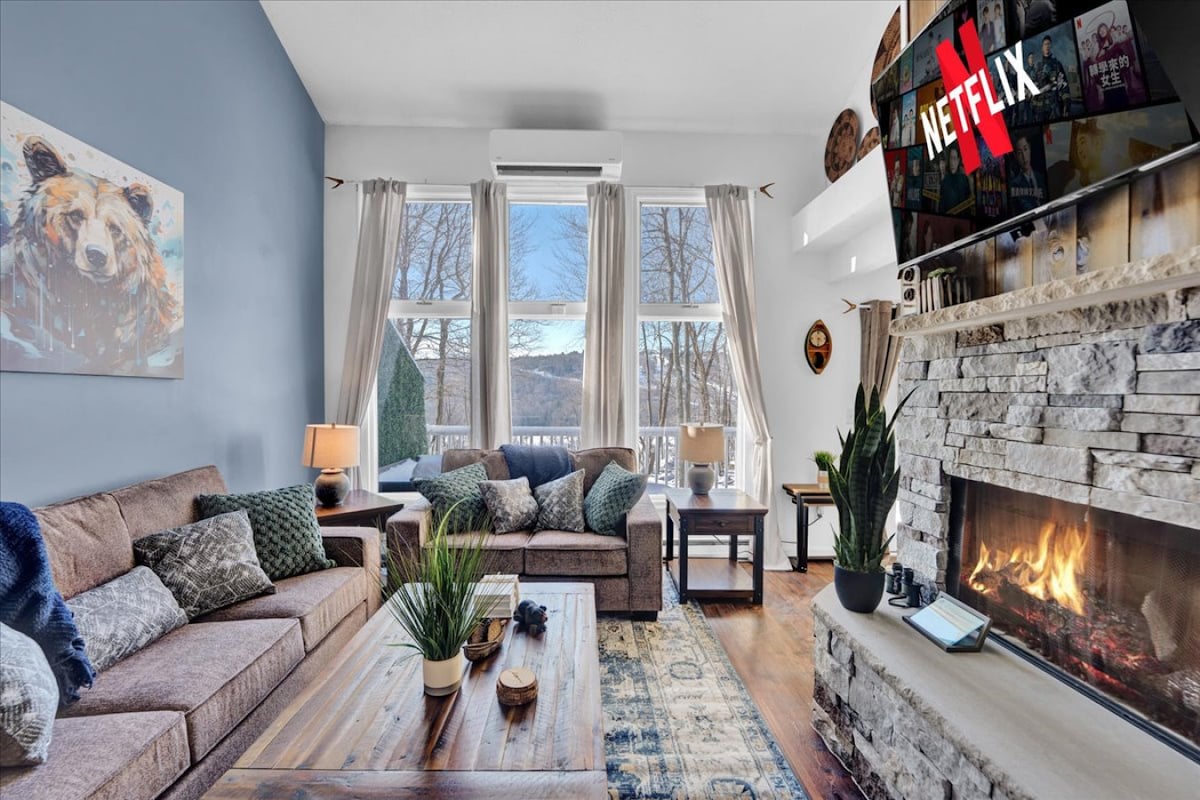 The living room features a spacious seating arrangement with plush couches and armchairs. A warm fireplace is present, alongside a large TV displaying streaming content. Natural light streams in through large windows, highlighting a tranquil view of the surrounding trees and mountains.