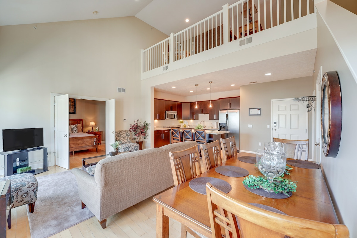 An open living area showcases a dining table set for six, accompanied by a cozy couch and armchair. The kitchen offers modern appliances and wooden cabinetry. A staircase leads to the upper level, while large windows allow natural light to fill the space.