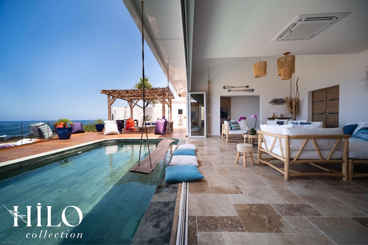 An open living space features a glass door that seamlessly connects to a wooden terrace and pool area. A suspended hammock and lounge seating are positioned by the pool. Natural stone tiles add texture, while vibrant plants provide a touch of greenery, enhancing the coastal ambiance.