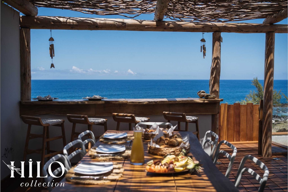 The dining area features a long wooden table set for a meal, overlooking the ocean. Natural light floods the space through an open structure made of woven branches. Bar stools enhance the casual atmosphere while the sea view creates a serene backdrop.