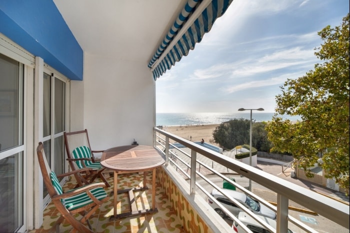 A spacious balcony features a round table and two striped chairs, offering a view of the sea and sandy beach. A green and white awning provides shade, and the floor is decorated with patterned tiles. The surrounding area includes lush greenery.