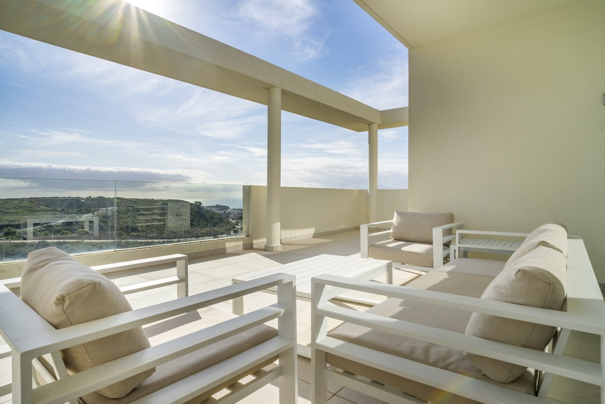 A spacious terrace is furnished with white outdoor seating, offering clear views of the surrounding landscape. Natural light enhances the clean lines of the furniture, while large columns and a glass railing frame the space, providing an open and airy feel.