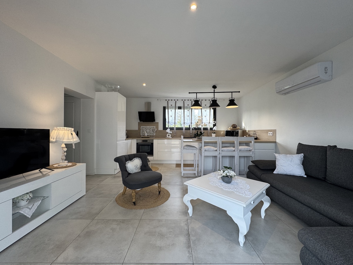 The spacious living area is illuminated by natural light, featuring a modern sofa and a cozy armchair. An adjoining fully equipped kitchen includes a dining bar with high stools. A decorative white coffee table adds elegance, while neutral tones create a calming ambiance.