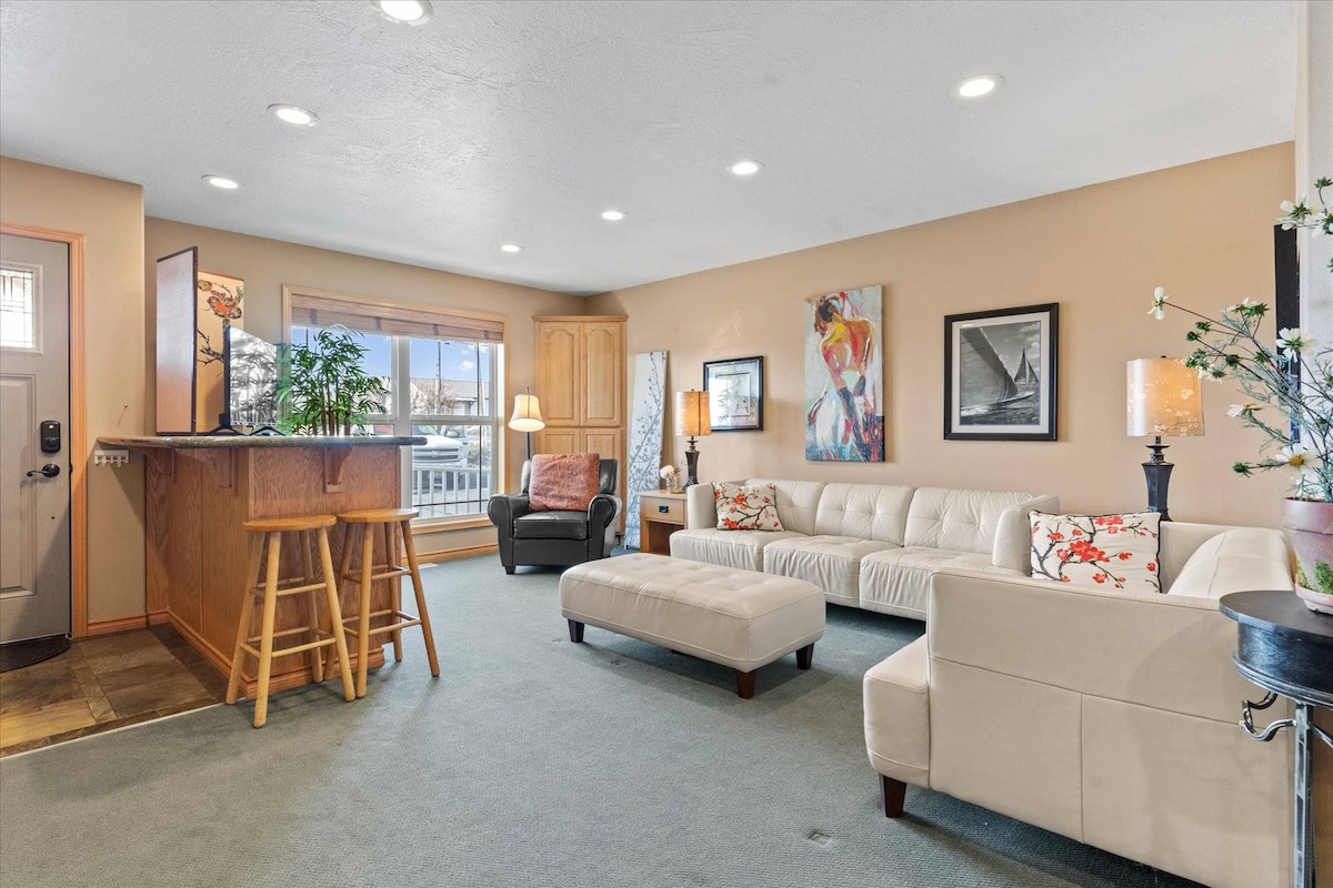 An open-concept living space features a light-toned sectional sofa, a patterned accent chair, and a coffee table. A kitchenette bar with stools is visible, along with artwork adorning the walls. Natural light floods in through a large window, enhancing the inviting atmosphere.