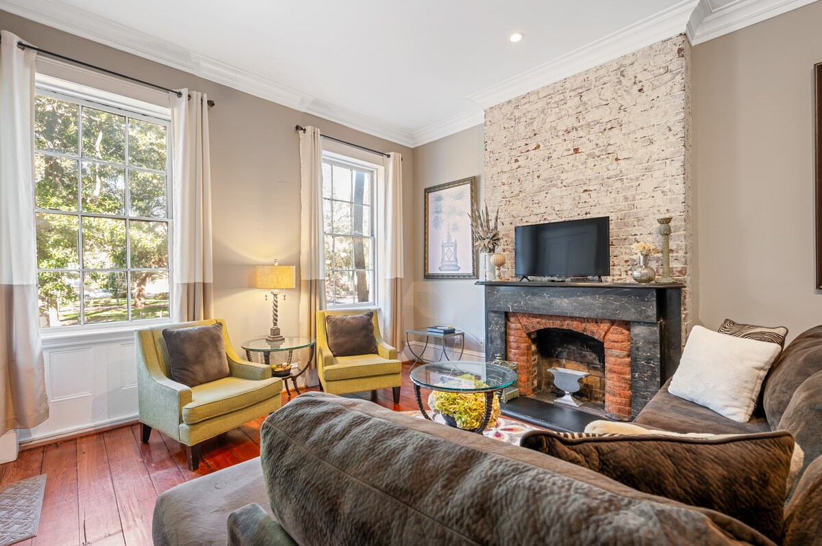 A living area features exposed brick walls and a decorative fireplace. Two green armchairs are positioned beside a side table, creating a cozy seating arrangement. Natural light floods in through large windows adorned with flowing curtains, enhancing the inviting ambiance.