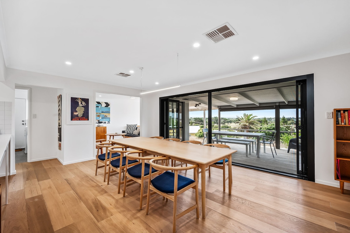 A spacious dining area is highlighted by a long wooden table surrounded by eight blue-accented chairs. Large glass doors open to an outdoor deck, providing views of the surrounding landscape, while warm wooden flooring adds a cozy touch to the interior.