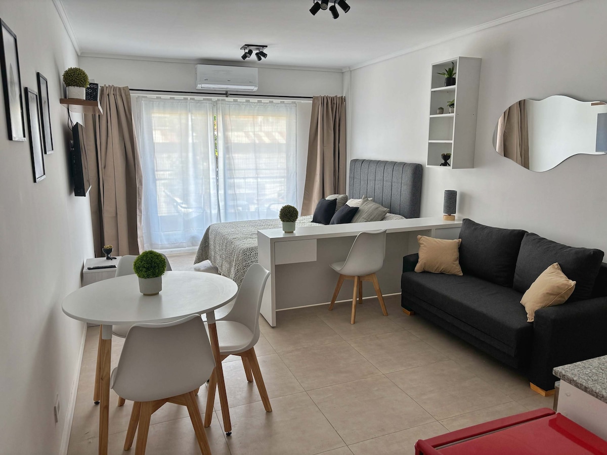 A modern studio layout is presented, featuring a spacious bed with a decorative headboard. A seating area with a dark couch is complemented by a small round dining table, surrounded by white chairs. Natural light filters through sheer curtains covering a large window.