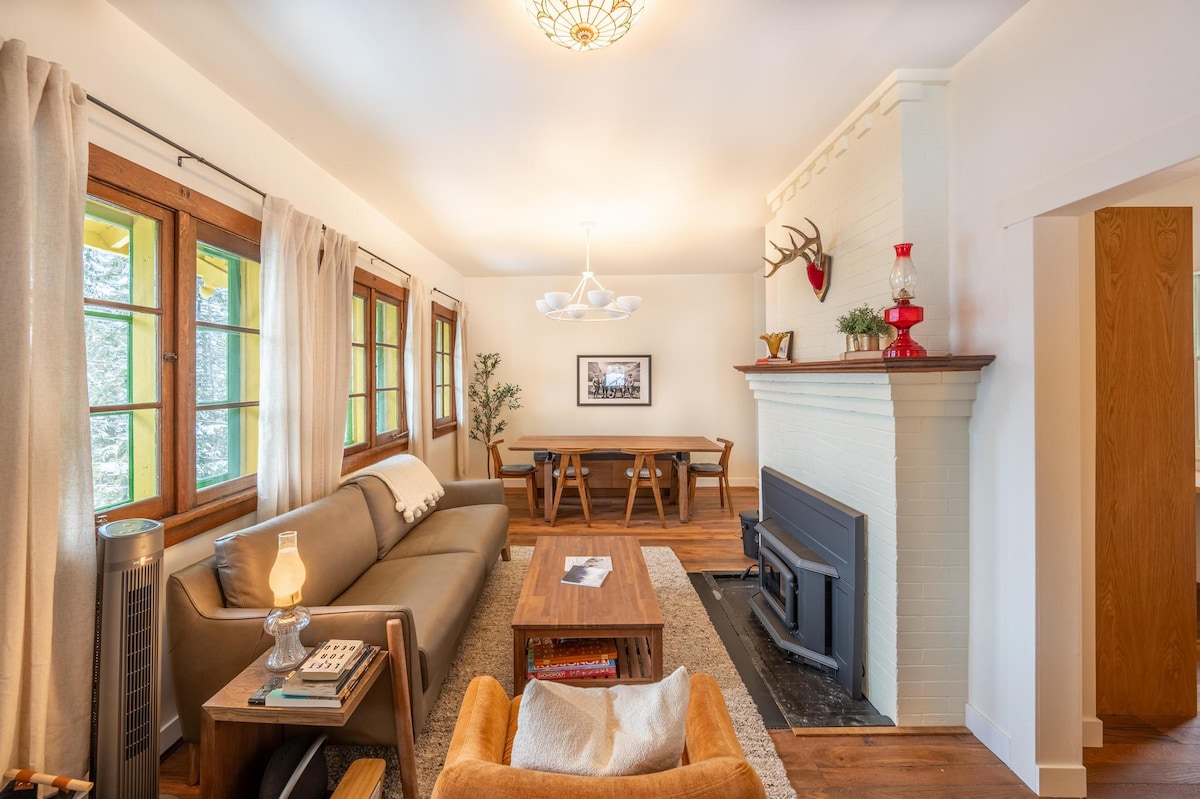 A spacious living area is highlighted by cozy seating arrangements and a warm fireplace. Large windows provide natural light and views of the surrounding landscape. A dining table is positioned nearby, complementing the inviting atmosphere of the chalet.