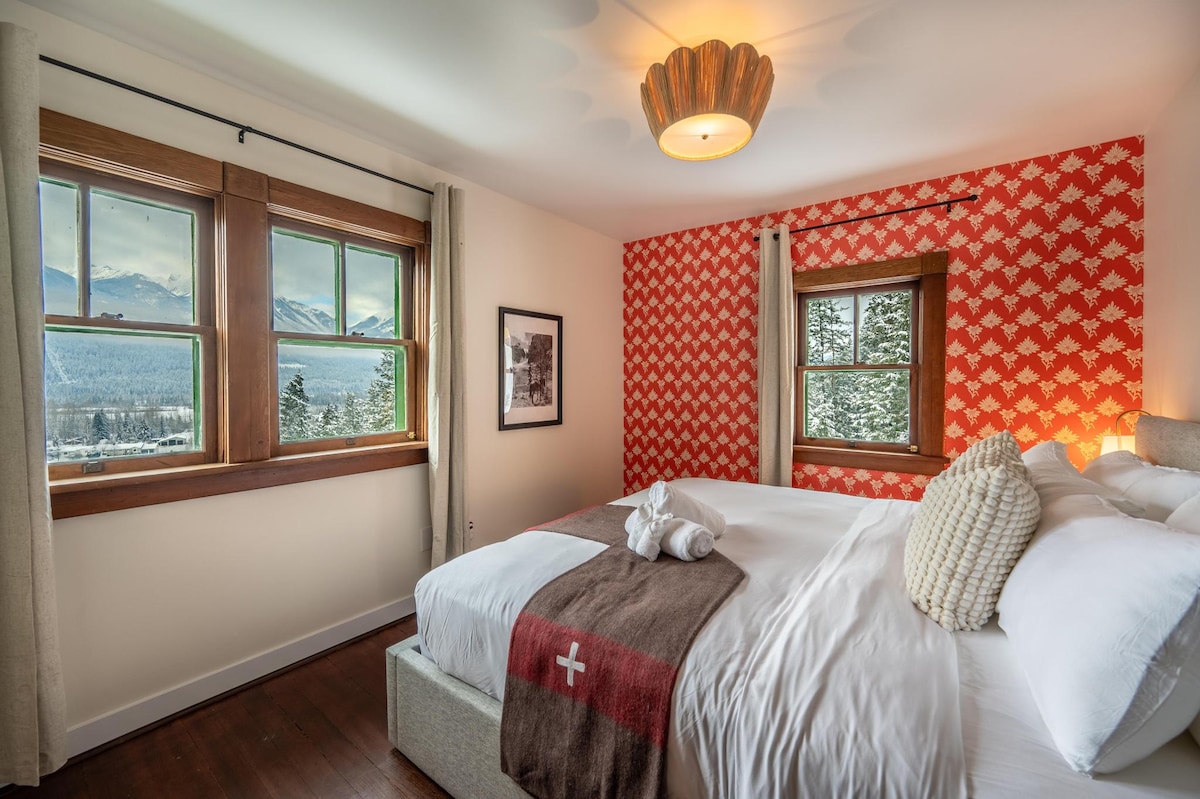 A serene bedroom space features a spacious bed adorned with a cozy blanket displaying a Swiss cross. The walls are decorated in a bold red motif, complemented by natural light streaming through two large windows, offering views of the surrounding mountains and snow-covered landscape.