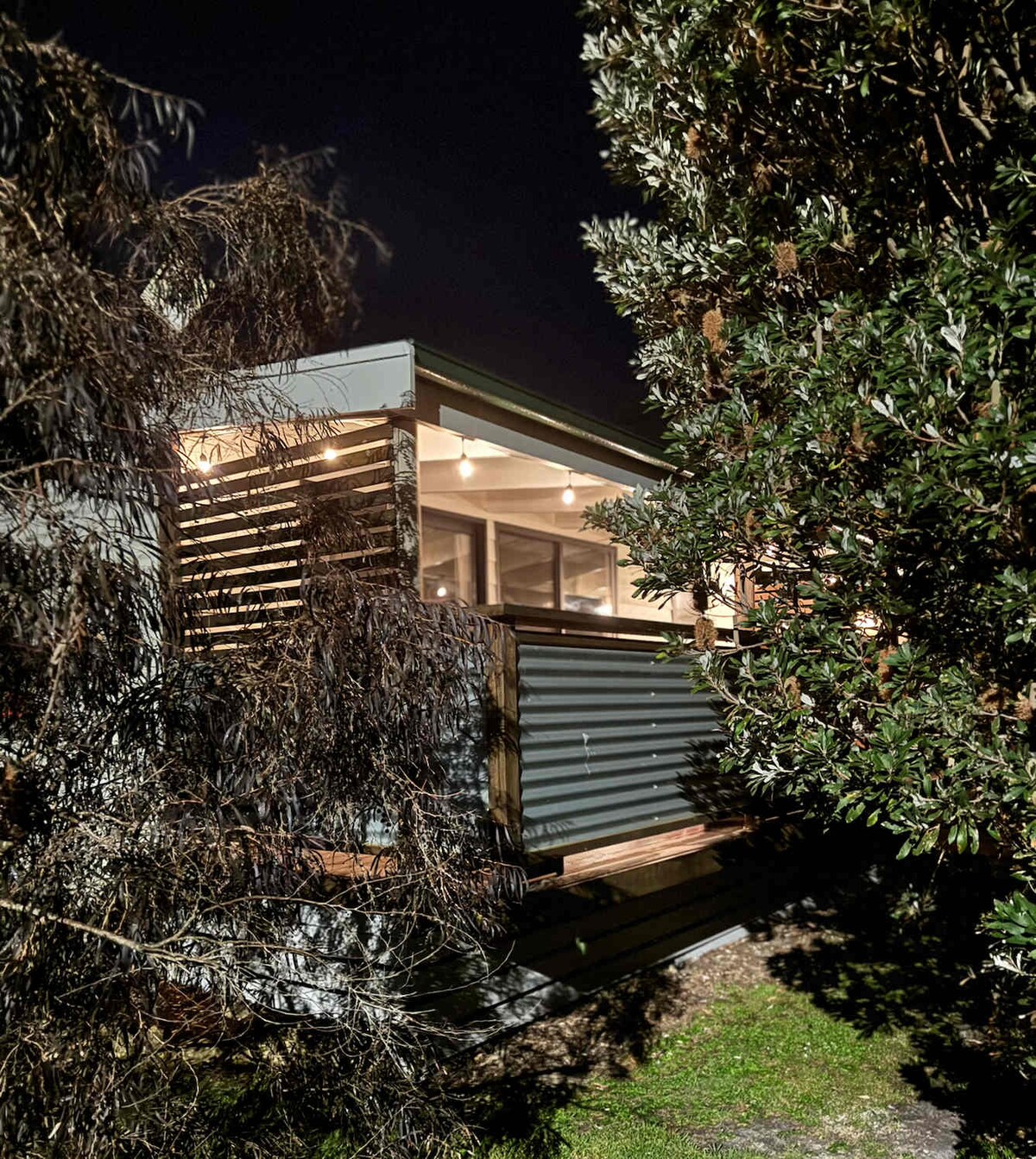 The exterior of the cottage is partially obscured by native vegetation, with warm lighting visible through the large windows. The balcony features wooden slats that provide privacy while maintaining an inviting appearance.
