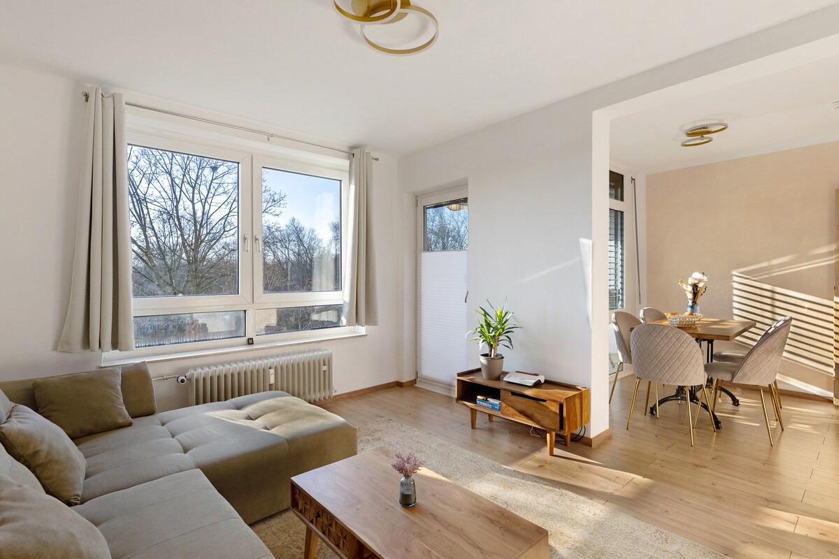 A modern living area is presented with large windows allowing natural light to fill the space. A comfortable sofa bed faces a wooden coffee table, and a dining area with a round table and four matching chairs is visible in the background.