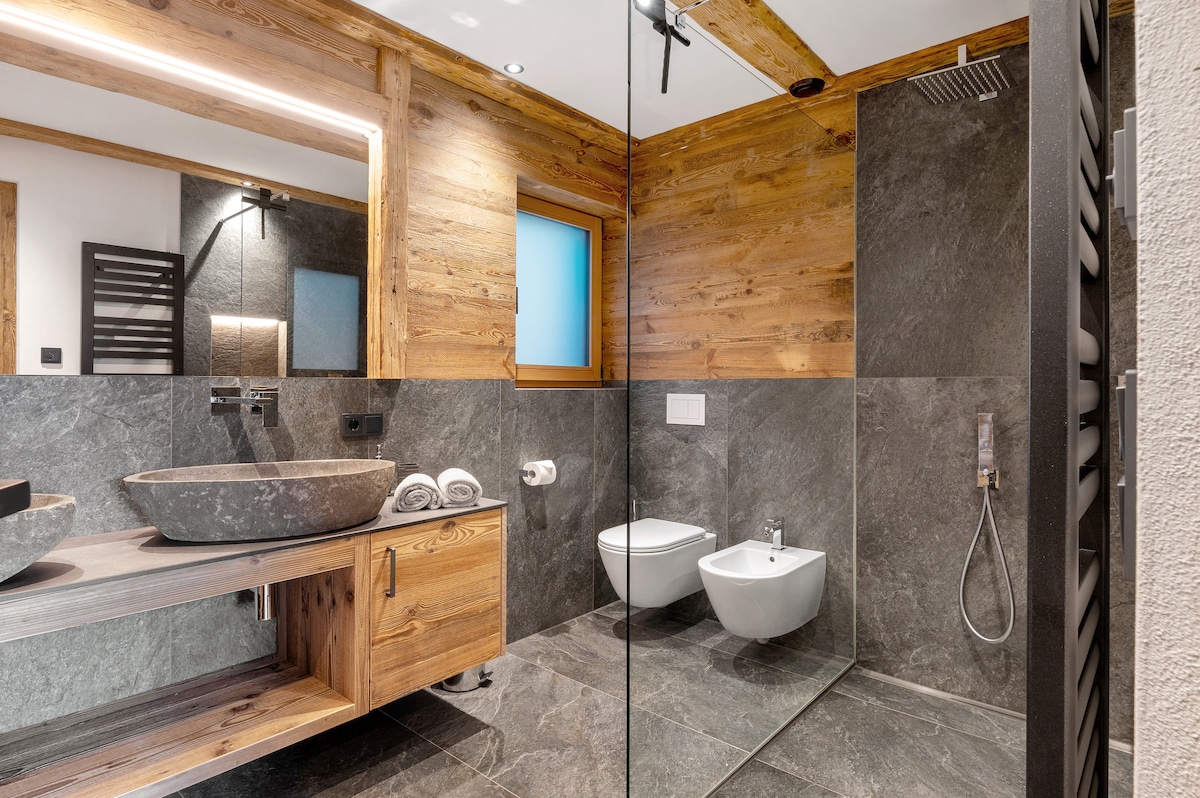 A modern bathroom features natural wood accents and stone-like surfaces. A circular sink is positioned on a wooden vanity, complemented by a large mirror. A walk-in shower with glass panels and minimalistic fixtures is visible. Two functional sanitary fixtures are placed beside the shower.