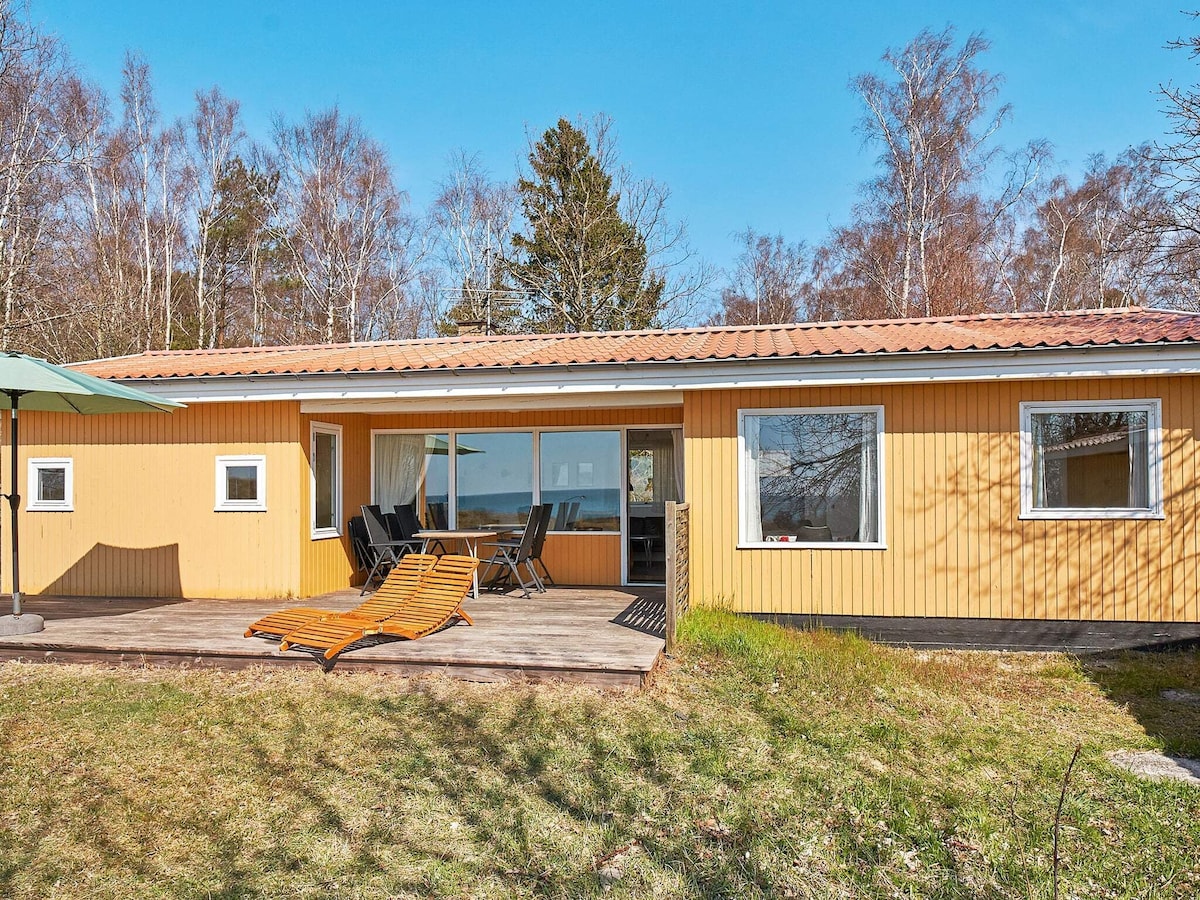 A holiday home with a yellow exterior features a spacious terrace, furnished with several black lounge chairs and sun loungers. Large glass sliding doors provide views of the surrounding garden and natural greenery. The setting is bright and open, highlighted by clear blue skies.