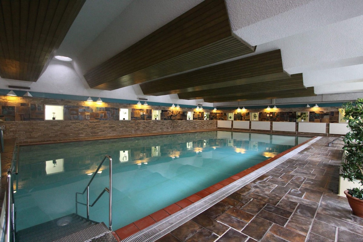 An indoor heated swimming pool is shown, featuring clear water and a smooth stone floor. The pool area is illuminated by soft overhead lighting, with natural stone accents around the walls. A decorative plant is positioned near the pool's edge.