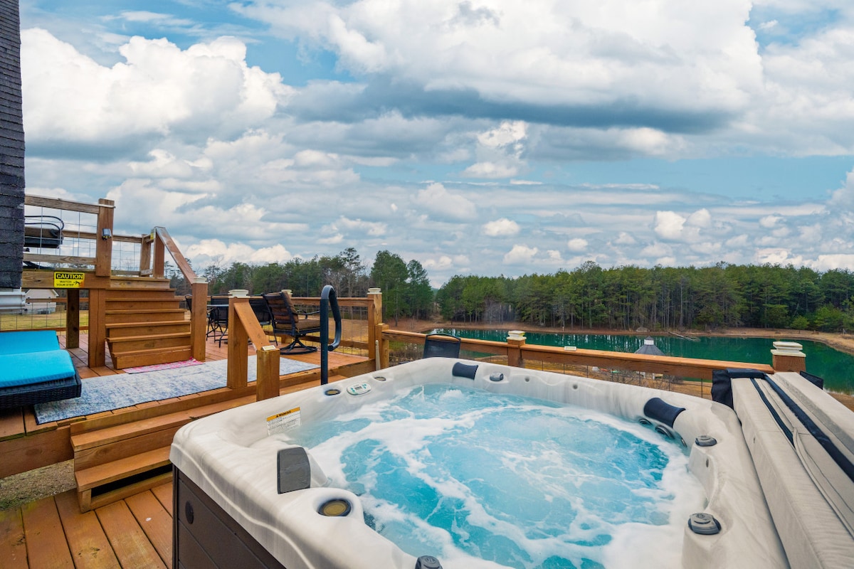 A private hot tub is positioned on a wooden deck, with water rippling in the foreground. A serene pond and lush trees are visible in the background, under a sky filled with clouds. Comfortable seating and steps leading from the deck complete the inviting outdoor scene.