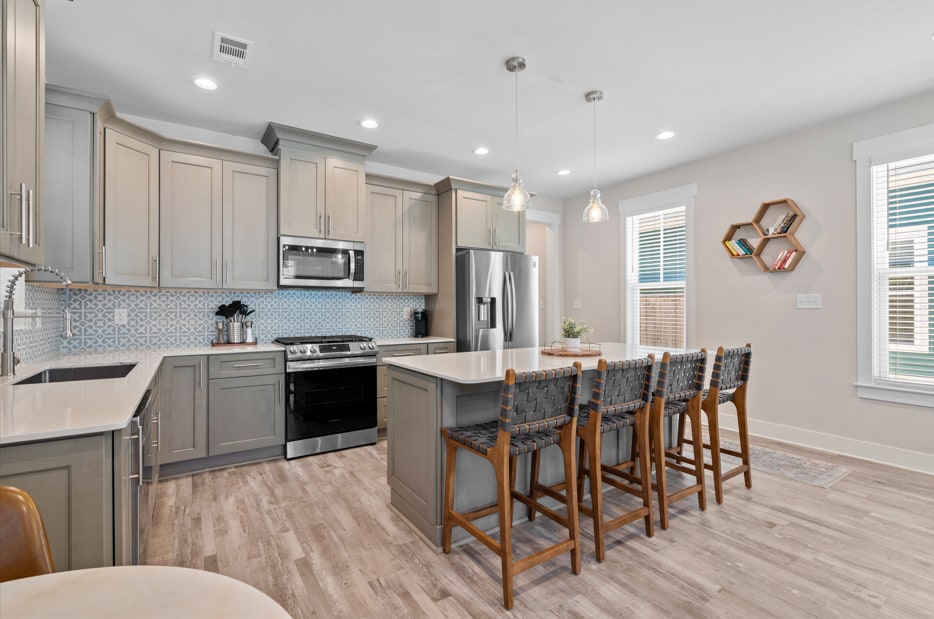 A modern kitchen features light grey cabinetry and stainless steel appliances. An island with bar seating for four is illuminated by pendant lights. Subtle blue tiles provide a patterned backsplash, while natural light filters in through nearby windows.