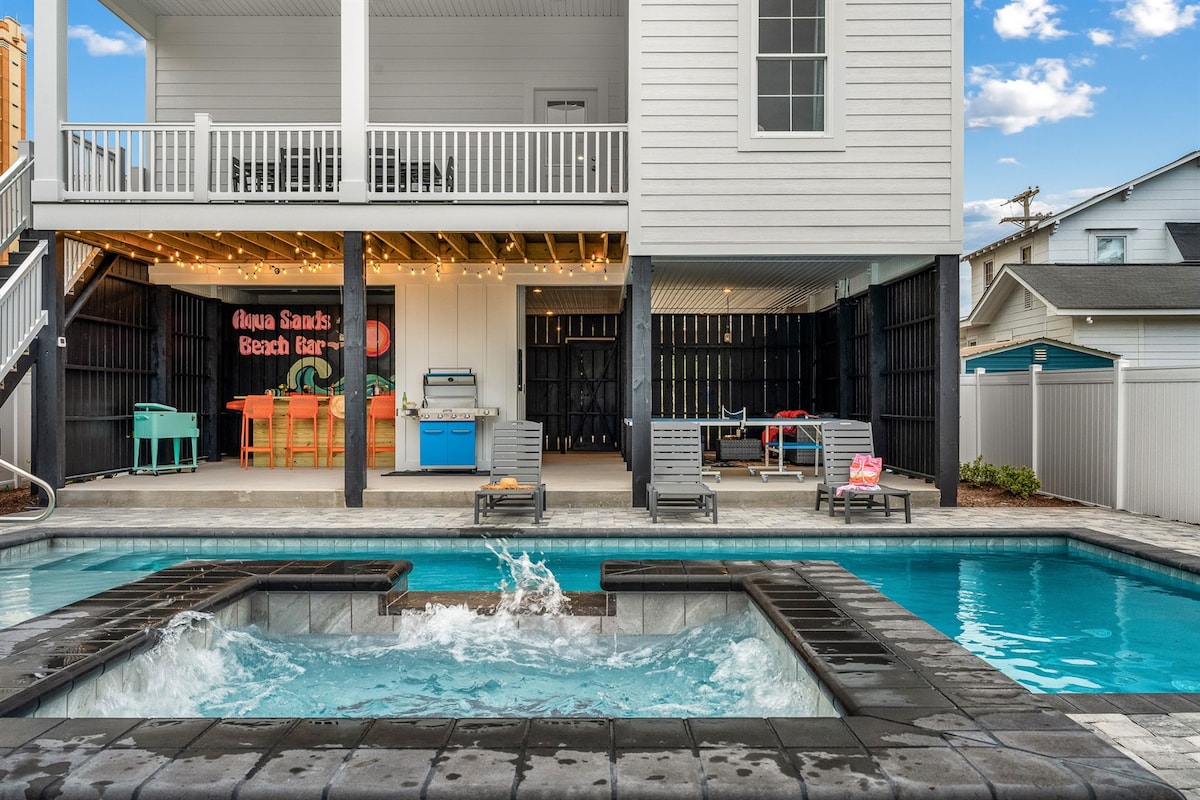 A private heated pool and spa are showcased in the inviting outdoor space, with bubbling water reflecting sunlight. The vibrant Tiki bar features colorful seating and is surrounded by string lights. Ample lounge chairs are positioned nearby under the covered porch.