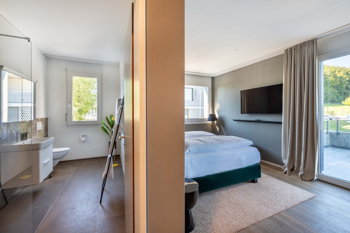 A spacious room is divided between a cozy sleeping area and a modern bathroom. Natural light enters through large windows, illuminating the bed with soft linens. A sleek flat-screen TV is mounted on the wall, and a plush rug is placed on the floor near the bed.
