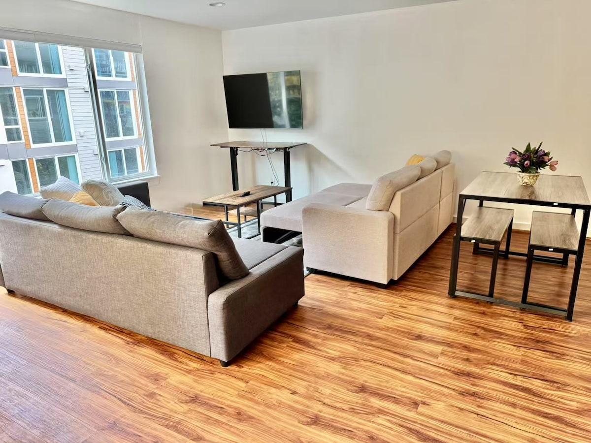 A spacious living area is highlighted by a large window allowing natural light to flow in. Two comfortable sofas face a wall-mounted TV, while a dining table with bench seating occupies one corner. The hardwood flooring complements the modern decor.