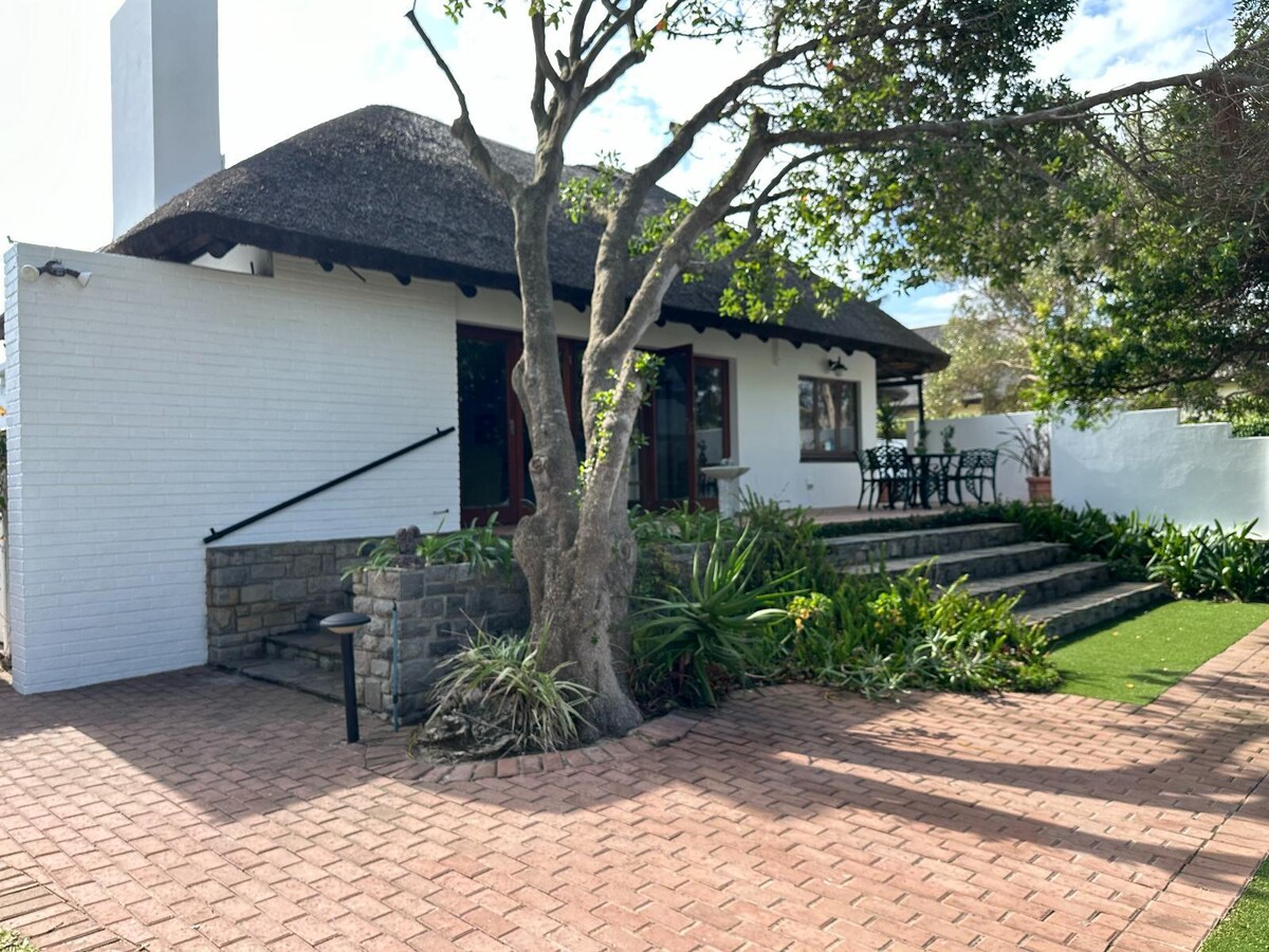 A charming exterior features a thatched roof and a stone pathway lined with greenery. Steps lead to a welcoming entrance, complemented by a small seating area in front. The well-maintained garden enhances the tranquility of the setting.