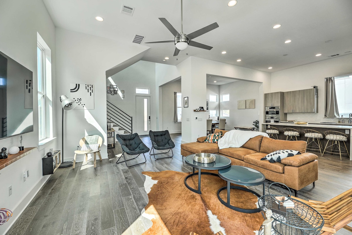 A modern living area displays an open layout featuring a large sectional sofa and two black lounge chairs. A round coffee table sits on a textured area rug. Large windows allow ample light to fill the space, while a ceiling fan provides ventilation above.