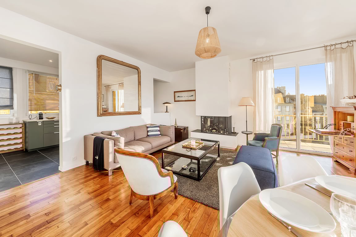 A bright living room is showcased with large glass doors leading to an outdoor terrace. A comfortable sofa, an armchair, and a coffee table are arranged on a textured rug. A dining area is visible with prepared table settings, and a stylish mirror reflects the room's inviting atmosphere.