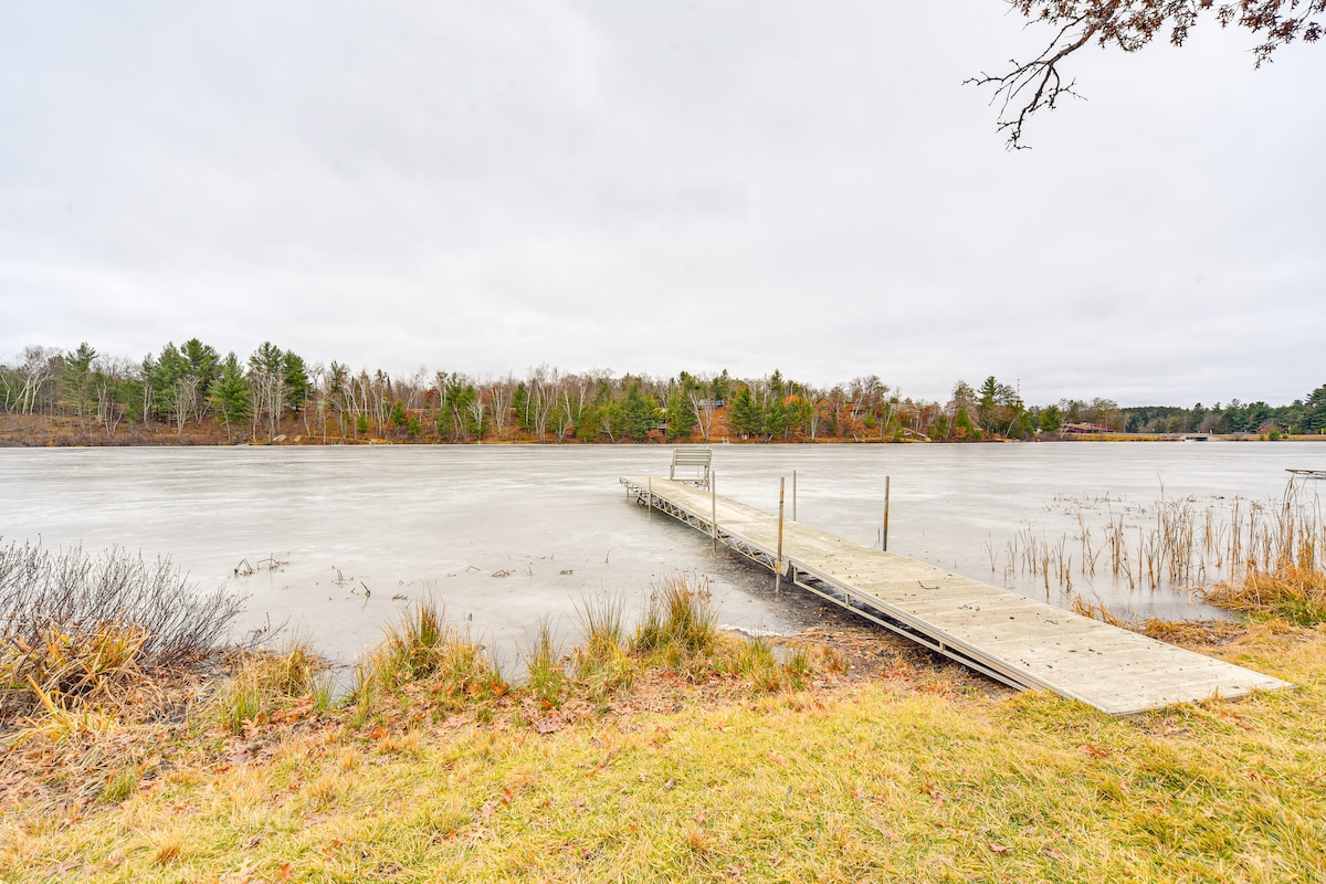 A wooden dock extends into a calm lake, surrounded by tall grasses and sparse trees. The shoreline is lined with earthy tones, and the water reflects an overcast sky, contributing to a tranquil atmosphere.