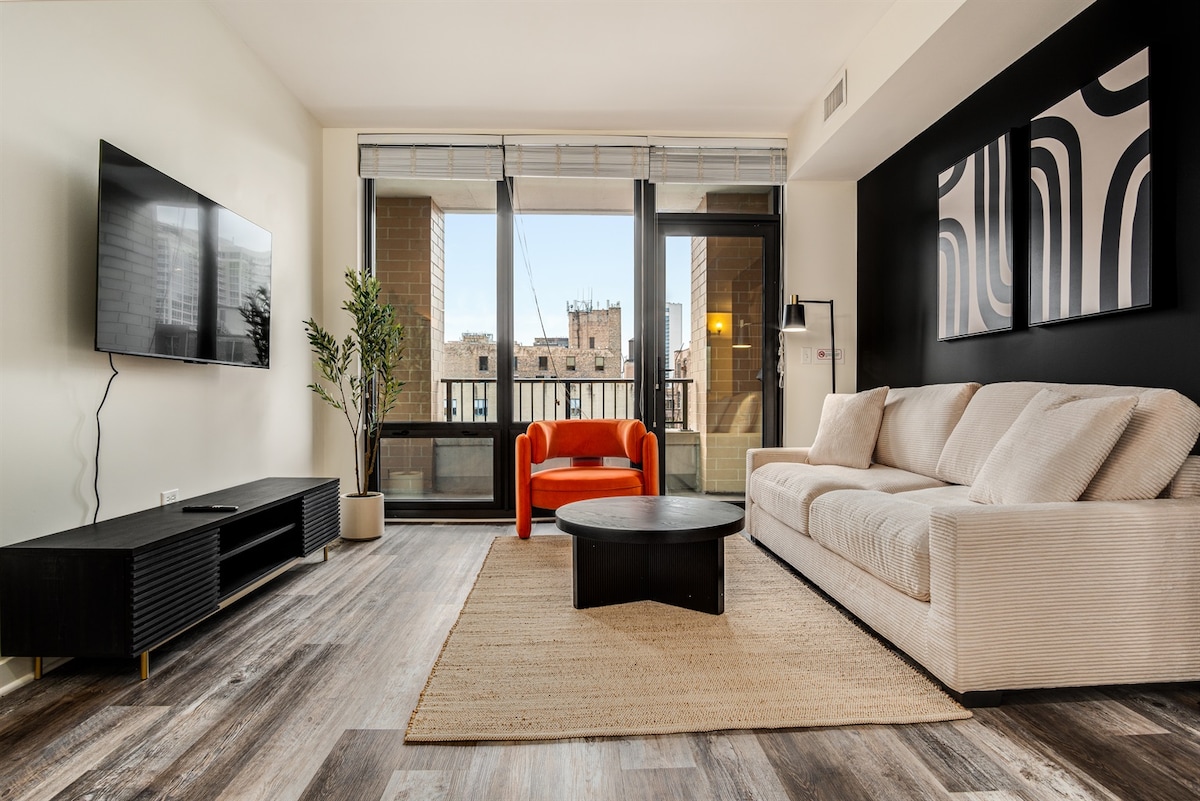A modern living area displays an open layout with a spacious beige sectional sofa and a striking red accent chair. Natural light fills the room through large glass windows, revealing a view of the city. A round black coffee table and a textured area rug add warmth to the space.