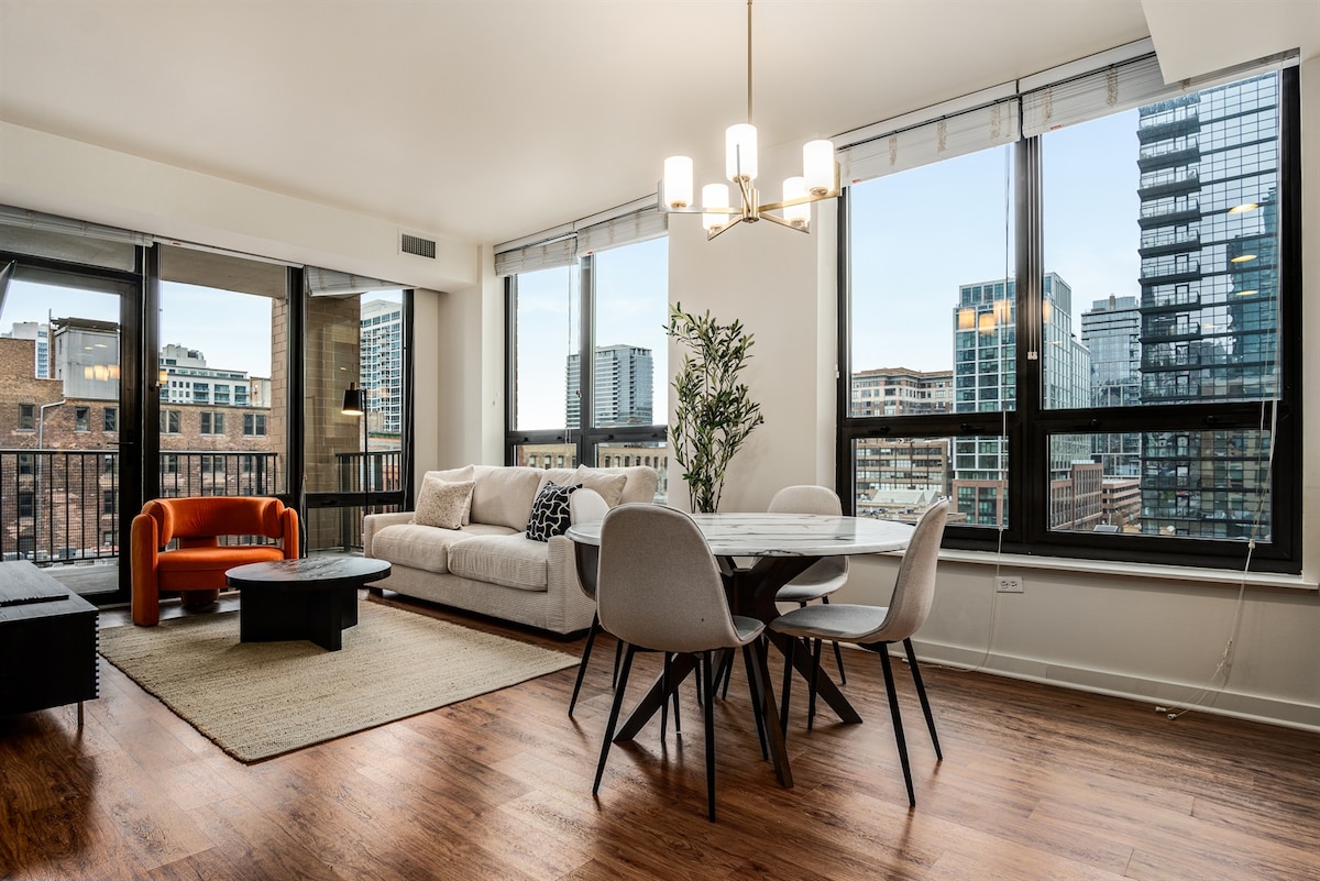 An open-concept living area features large windows that offer skyline views. A comfortable sofa and an orange accent chair are complemented by a modern dining table surrounded by stylish chairs. A neutral area rug adds warmth to the hardwood floor.