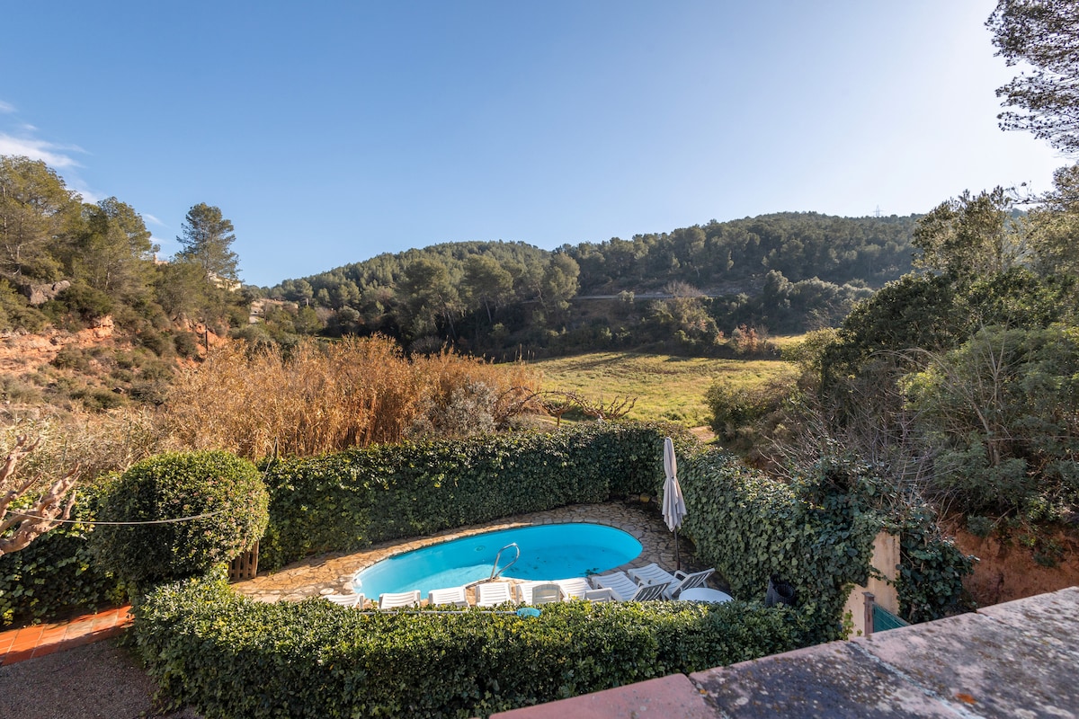 A pool is situated within a stone-enclosed area, surrounded by lush greenery and tall trees. Sun loungers are positioned nearby, providing a place for relaxation. The view features rolling hills and natural scenery under a clear blue sky.