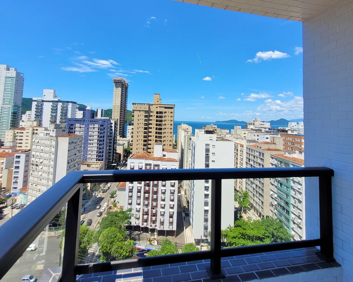 A panoramic view captures the vibrant cityscape and distant blue sea. High-rise buildings line the streets below, while a clear sky with scattered clouds stretches overhead. A balcony railing frames the scene, inviting guests to enjoy the refreshing atmosphere.