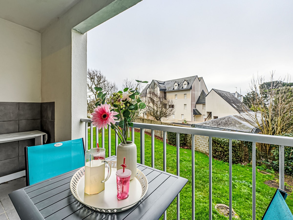 A balcony setting is highlighted, featuring a table with a pitcher and two glasses in vibrant colors. A vase with flowers adds a touch of nature. The view includes a grassy area and buildings in the distance, creating a serene outdoor ambiance.