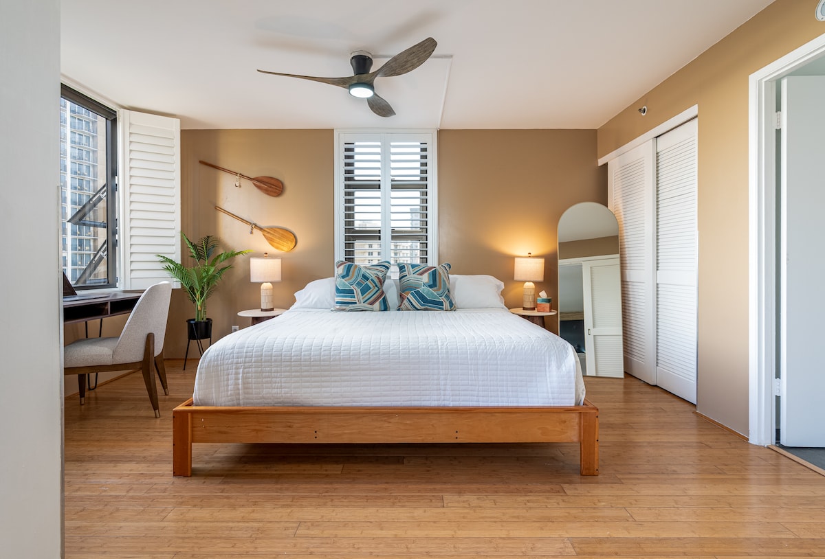 A spacious bedroom features a king bed with decorative pillows, complemented by bamboo flooring. Natural light filters through two windows, highlighting a stylish desk and chair. A full-length mirror and wall-mounted shelving enhance the modern design, creating a comfortable and inviting atmosphere.