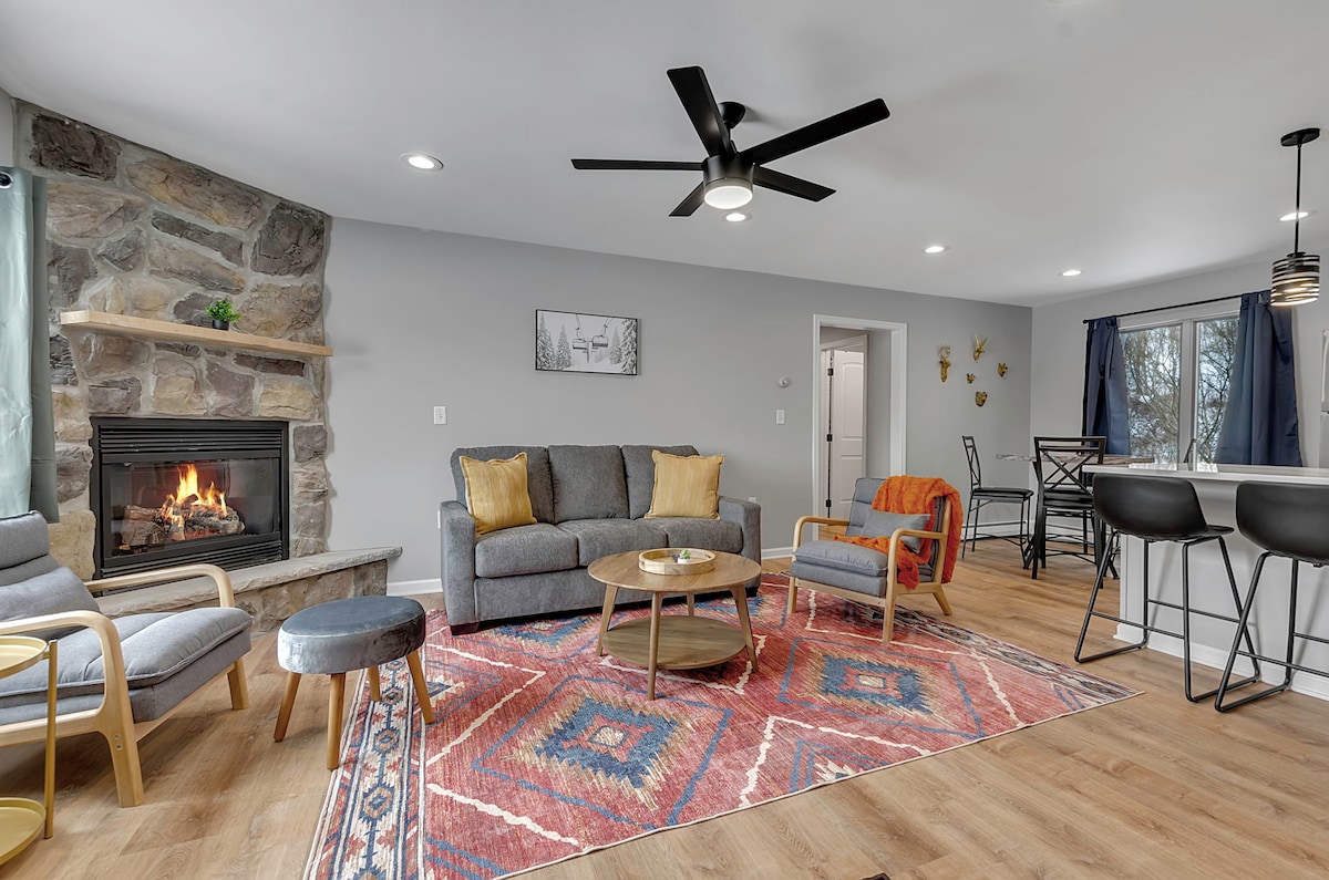 A spacious living area is highlighted by a stone fireplace, providing a focal point. Comfortable seating, including a sofa and armchairs, is arranged around a coffee table. Decorative elements include a patterned rug and large windows with simple curtains, allowing natural light to fill the room.