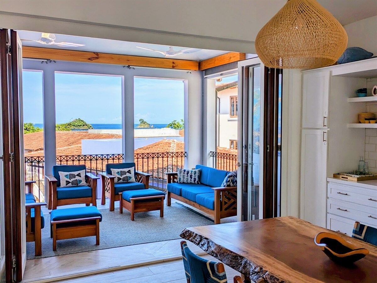 A bright living space features an open layout with large windows and doors offering ocean views. Comfortable seating is arranged on a light-colored rug, harmonizing with the wooden dining table. Natural light floods the area, enhancing the coastal ambiance.