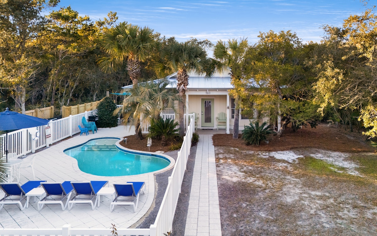 A spacious outdoor area features a private heated pool surrounded by sun loungers and tropical landscaping. A white fence encloses the space, providing privacy and serenity. The entrance to the house is visible, with inviting seating on the porch under the shade of palm trees.