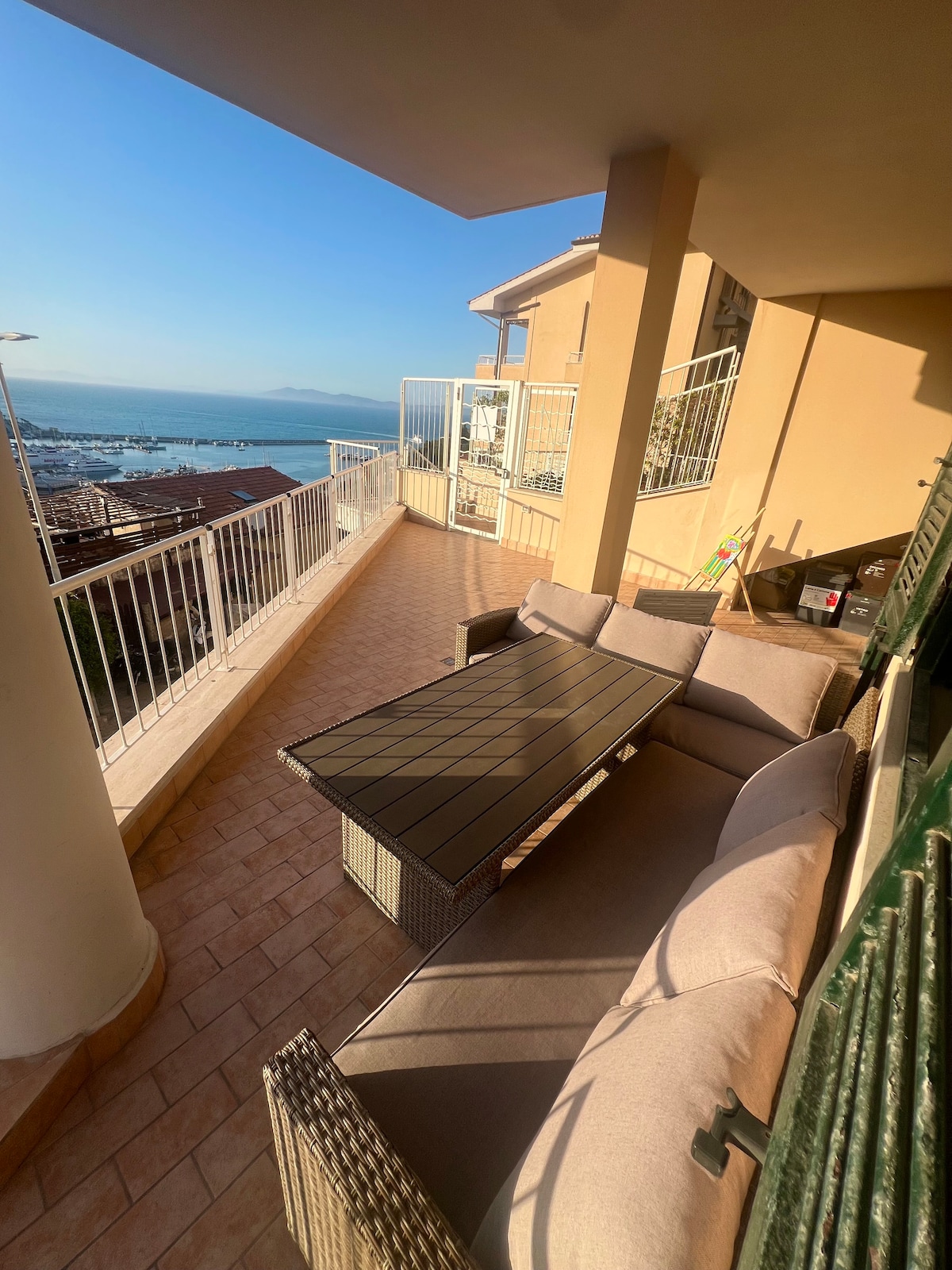 An outdoor terrace features a spacious seating area with a rectangular table and comfortable cushions. The railing offers a clear view of the surrounding area, including the bay and distant mountains under a clear blue sky.