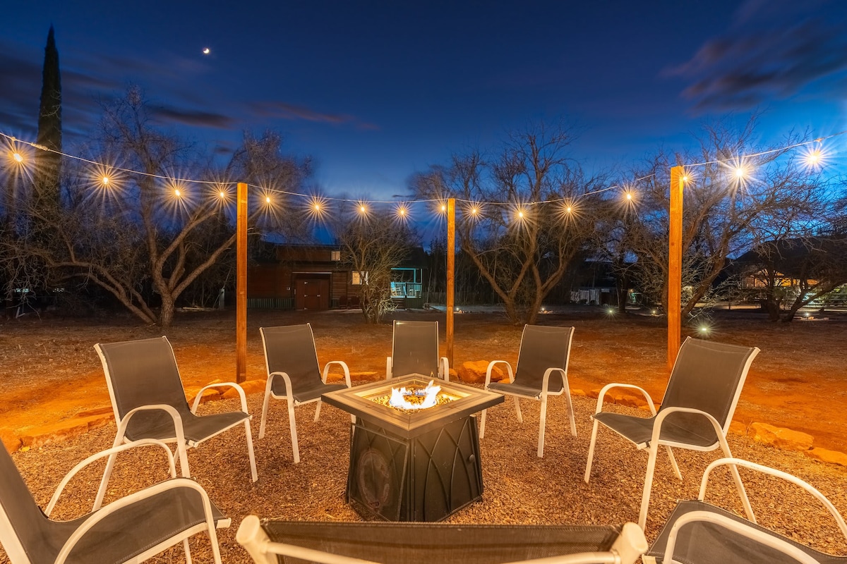 A shared fire pit lounge area is highlighted under a twilight sky. Six white lounge chairs surround a central fire pit, with string lights illuminating the space. The setting is bordered by sparse trees, providing a tranquil atmosphere for evening gatherings.