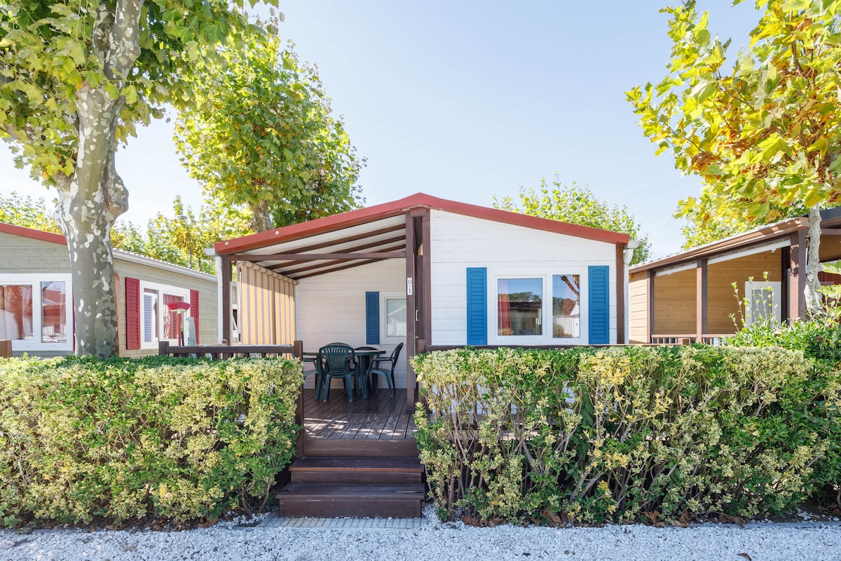 A bungalow features a covered porch with a small table and chairs, surrounded by neatly trimmed hedges. The exterior showcases a mix of light and dark wood, framed by vibrant green trees. Brightly colored shutters add a cheerful touch to the façade.