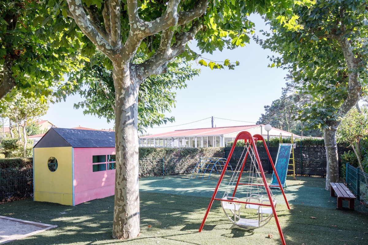 An outdoor children's play area is shown, featuring a colorful playhouse and a swing set. The ground is covered with artificial grass, surrounded by trees that provide shade. Bright sunlight creates a cheerful ambiance, inviting children to play in a secure environment.