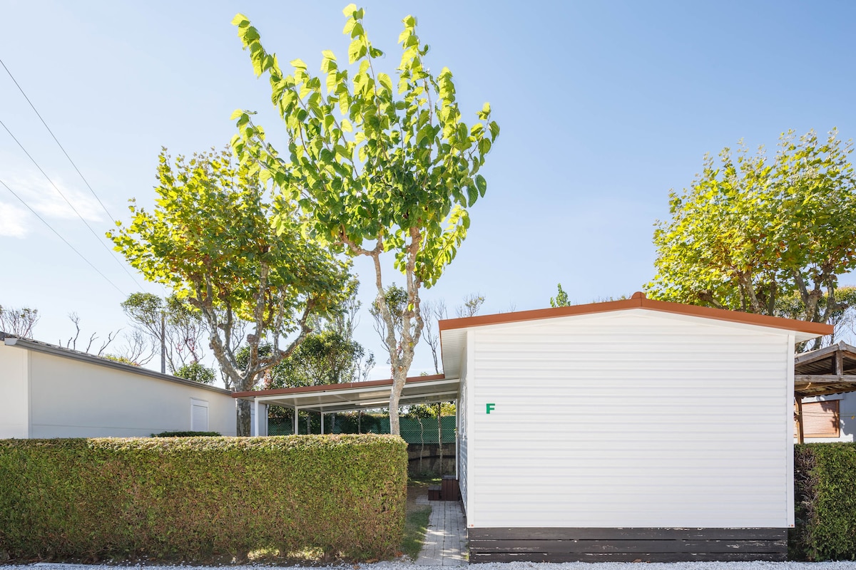 A bungalow with a clean, white exterior is surrounded by well-maintained greenery and trees. The entrance is sheltered under an overhang, providing shade. The space is designed for easy accessibility, with a serene outdoor setting indicated by the lush foliage.