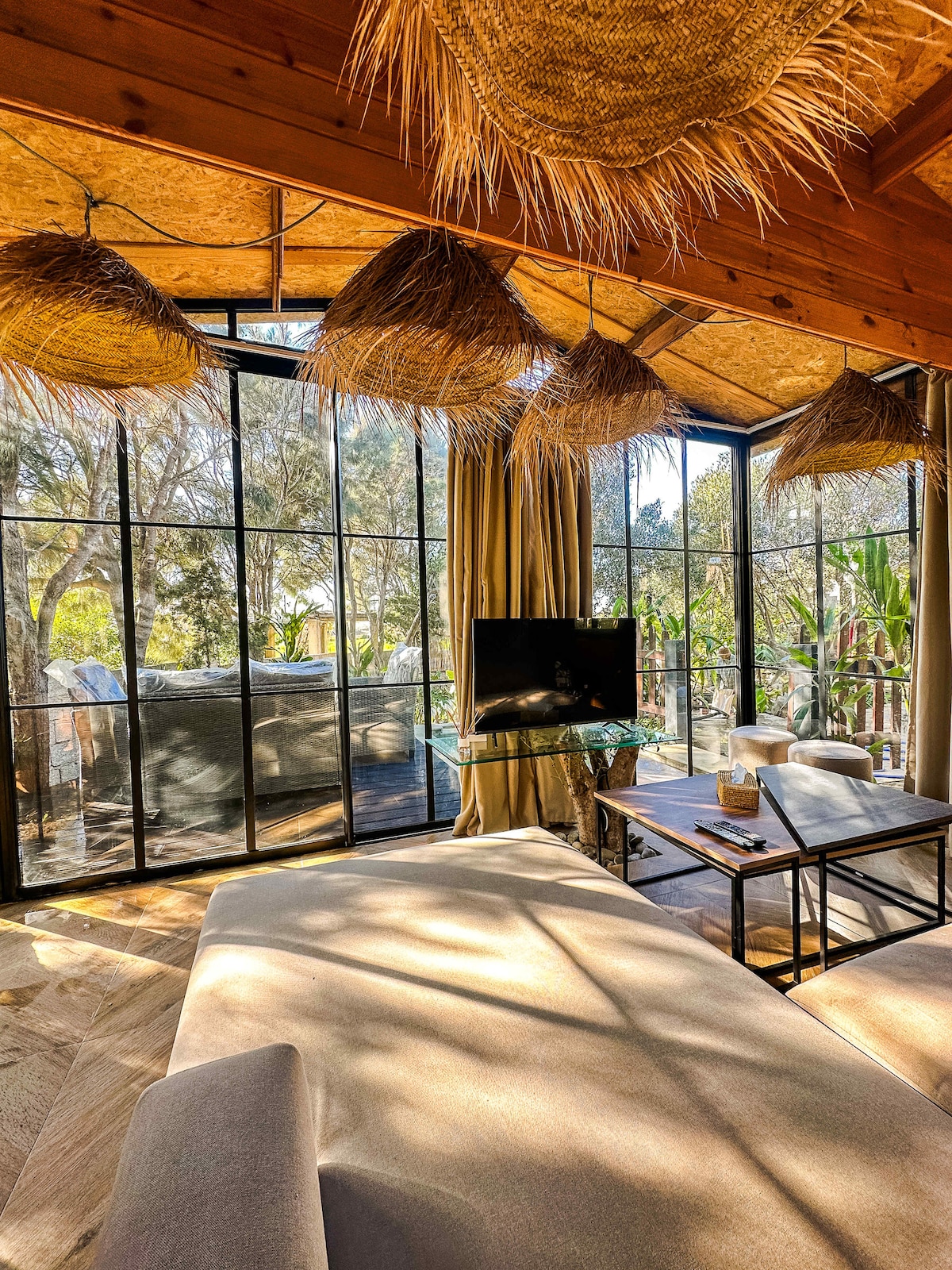 A spacious living area features large glass windows, allowing natural light to flood the space. Stylish straw light fixtures hang from the ceiling. A comfortable sofa and a wooden coffee table with decorative elements are arranged for relaxation, providing a view of the lush outdoor surroundings.