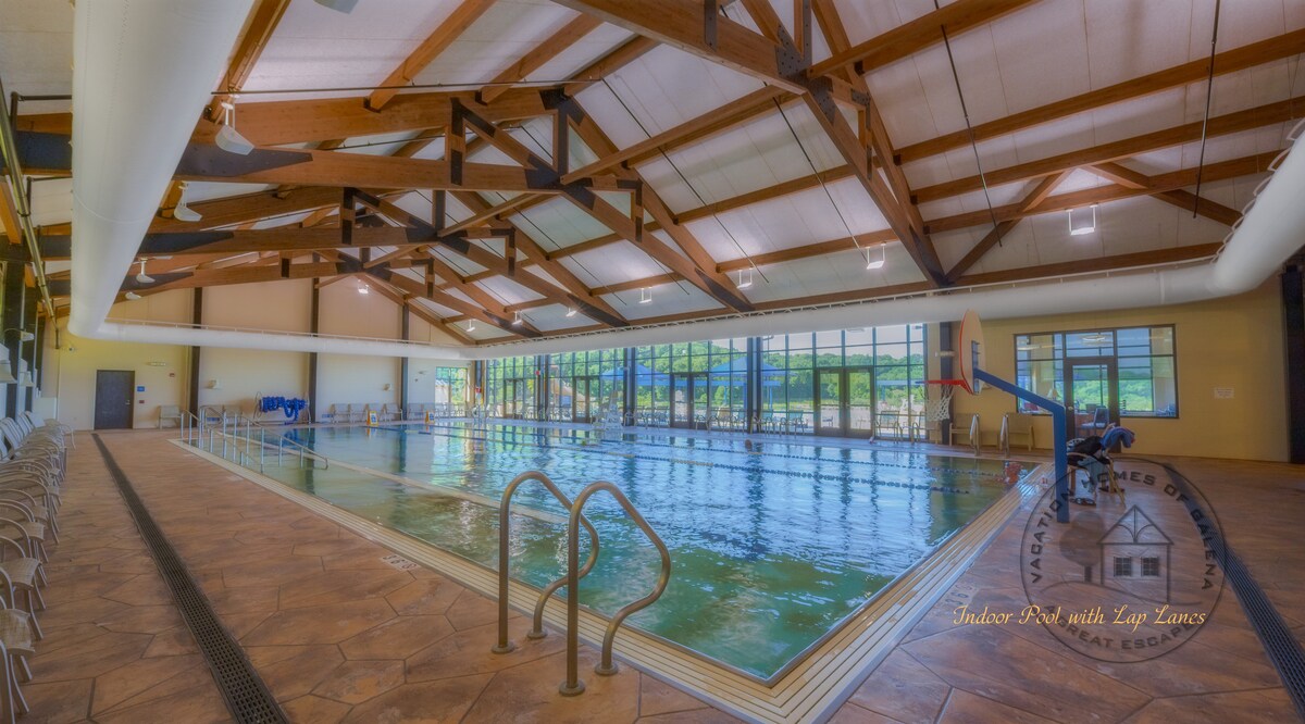 A spacious indoor pool area features large windows allowing natural light to fill the space. This facility includes multiple lap lanes, comfortable seating along the side, and a basketball hoop at one end. The ceiling showcases exposed wooden beams contributing to the open feel.