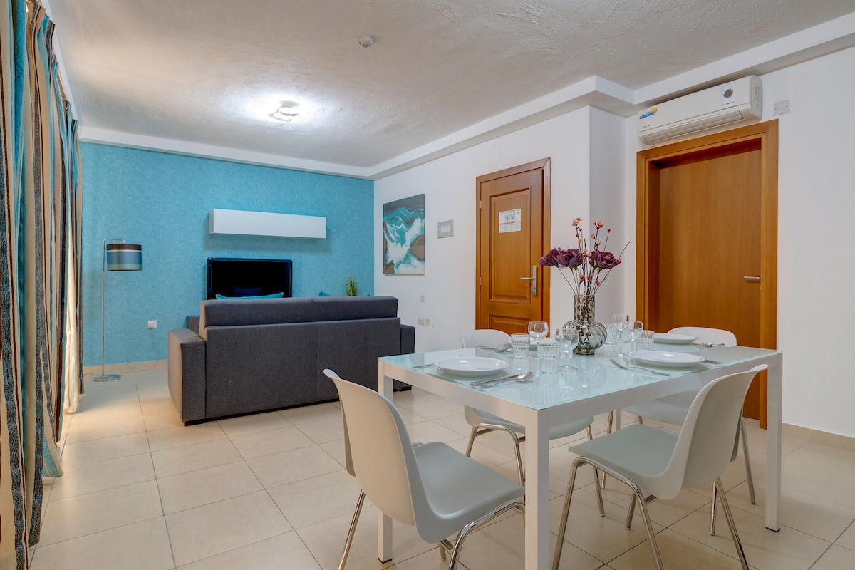 A modern living area is highlighted by a grey sofa and a dining table set for four. Light blue walls complement the neutral tones of the furniture. Decorative flowers are arranged in a glass vase, while an artwork adds a touch of character to the space.