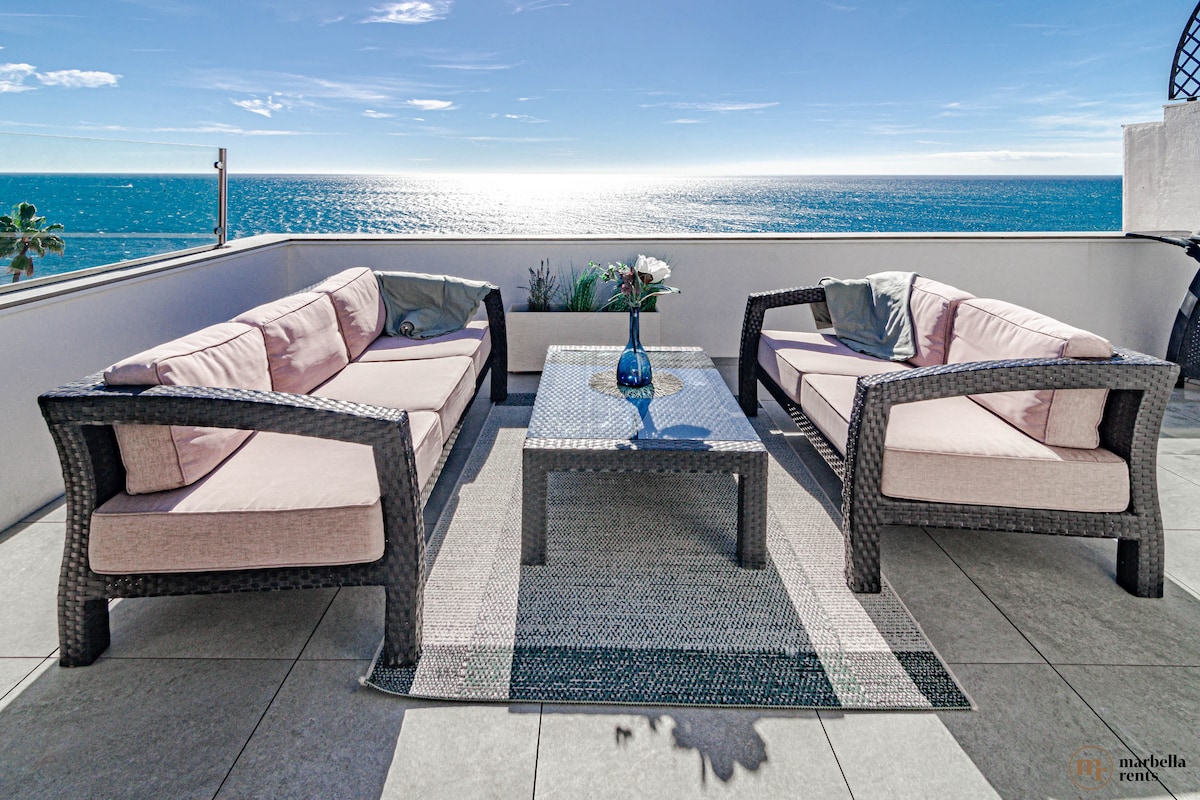 An outdoor terrace is shown with two cushioned seating areas facing the ocean. A glass coffee table sits between the seating, with a blue vase holding flowers. The backdrop features a clear blue sky meeting the serene sea, creating a relaxing atmosphere.