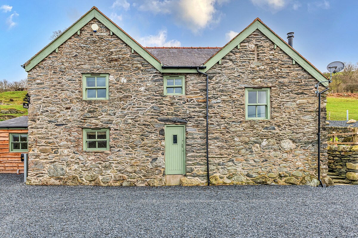 The stone exterior of Moelis Barn is prominently displayed, featuring a light green door and windows. The property sits on a gravel driveway, surrounded by a scenic hillside, showcasing the traditional farmhouse design.