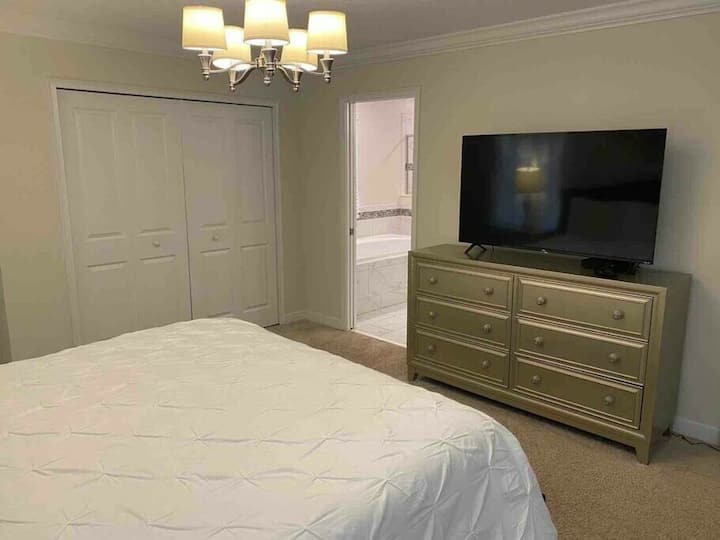 This cozy bedroom features a soft, neutral palette complemented by plush carpeting and a stylish dresser. A contemporary chandelier adds a touch of charm, while the large TV offers entertainment at your fingertips.