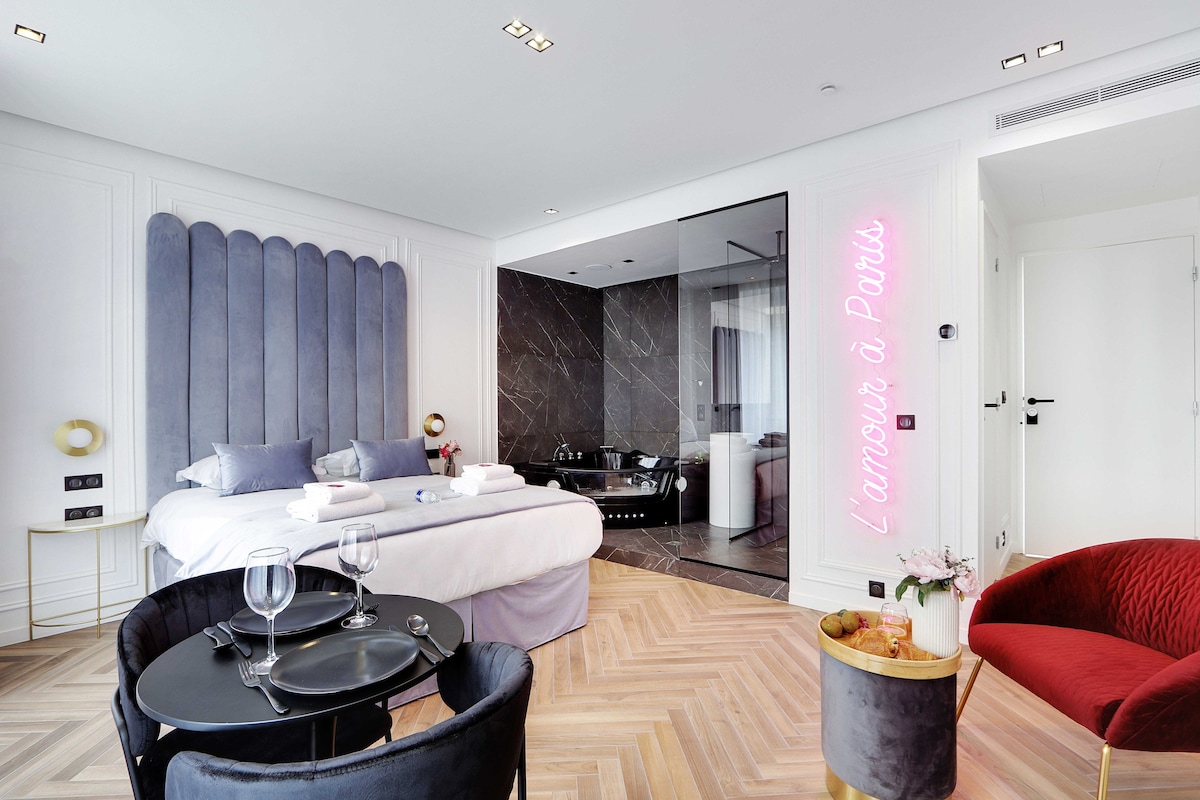 A modern suite features a queen size bed with blue accents and elegant decor. A dining area with a circular table and chairs complements the inviting atmosphere. The bathroom is visible through glass walls, showcasing a walk-in shower and a jacuzzi, highlighted by soft neon lighting.