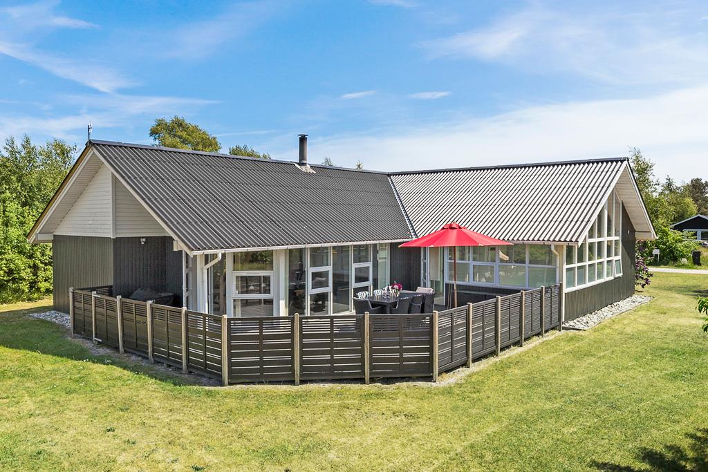 A modern holiday home is showcased with a sloped roof and large windows. A spacious terrace is visible, furnished with a dining set under an umbrella. The surrounding lawn is well-maintained, with greenery that enhances the outdoor space.